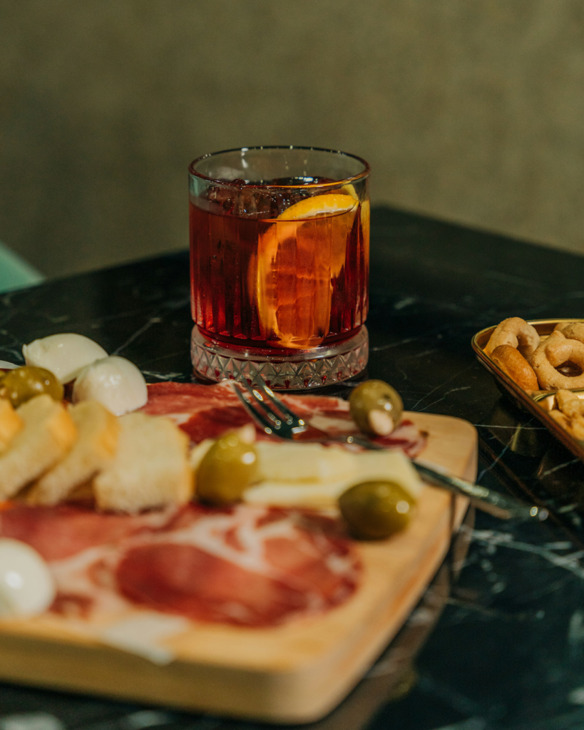 Negroni Cocktail mit Orangenschale neben italienischen Aperitivo Snacks wie Prosciutto, Käse und Oliven.