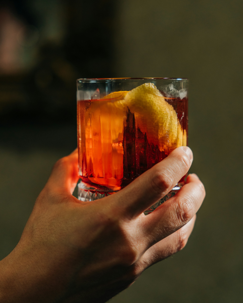 Hand hält einen klassischen Negroni Cocktail mit Orangenschale – ikonisches Getränk der italienischen Aperitivo-Kultur.